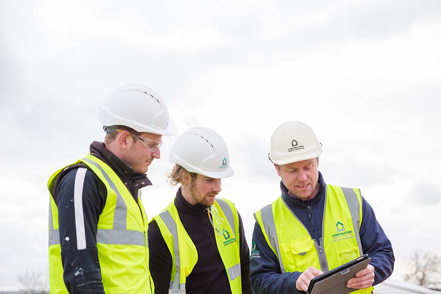 Three construction contractors on site in high-visibility jackets and hardhats, review their contractor management software on a tablet.